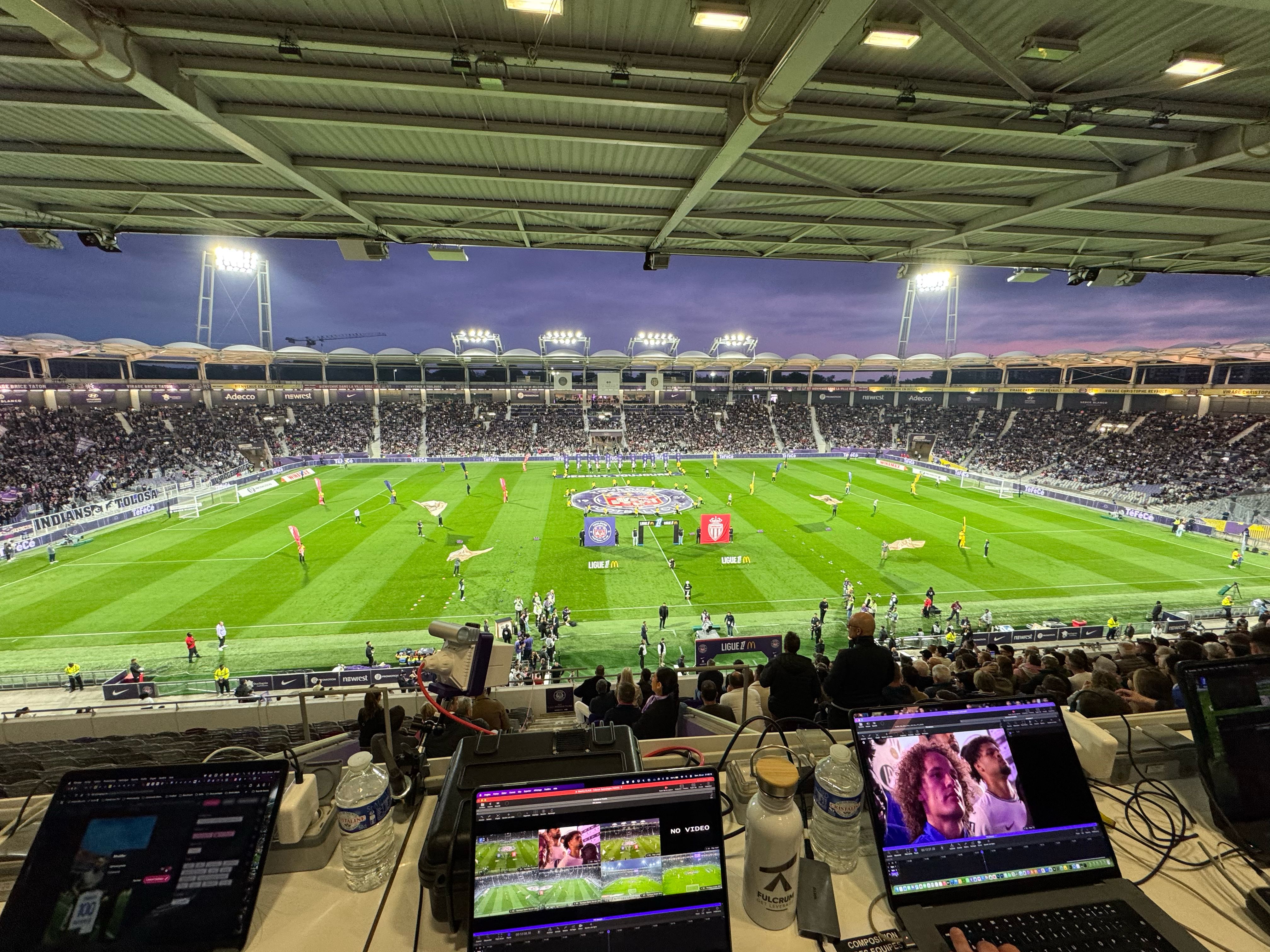 Laptops running analysis software on a desk high in the stand of a floodlit football stadium before kick-off, with a capture case and a camera at the front of the desk.
